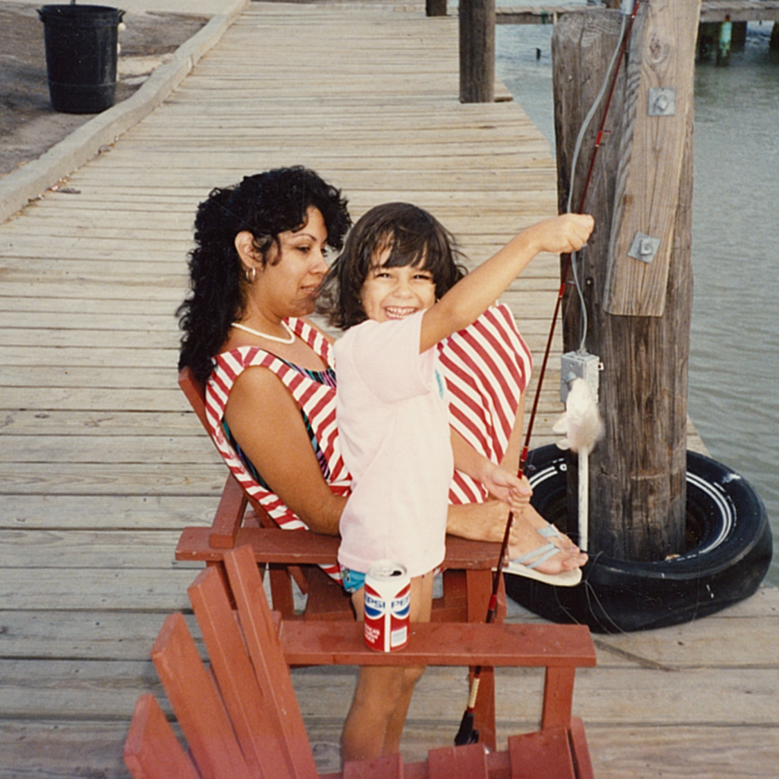 young girl sitting on her mother's lap on a dock holding a fishing pole and smiling at the camera.g 