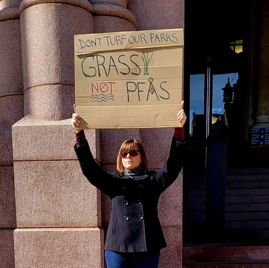 Cynthia holds a cardboard protest sign that reads, "Don't Turf Our Parks, Grass Not PFAS"