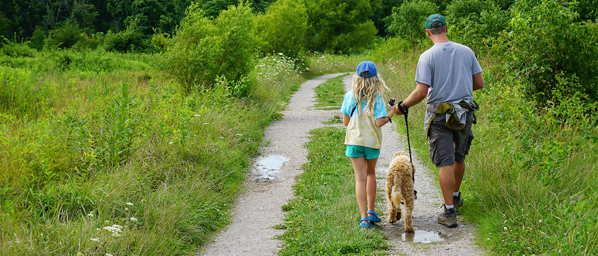 A father and daughter walk down a trail with their dog.