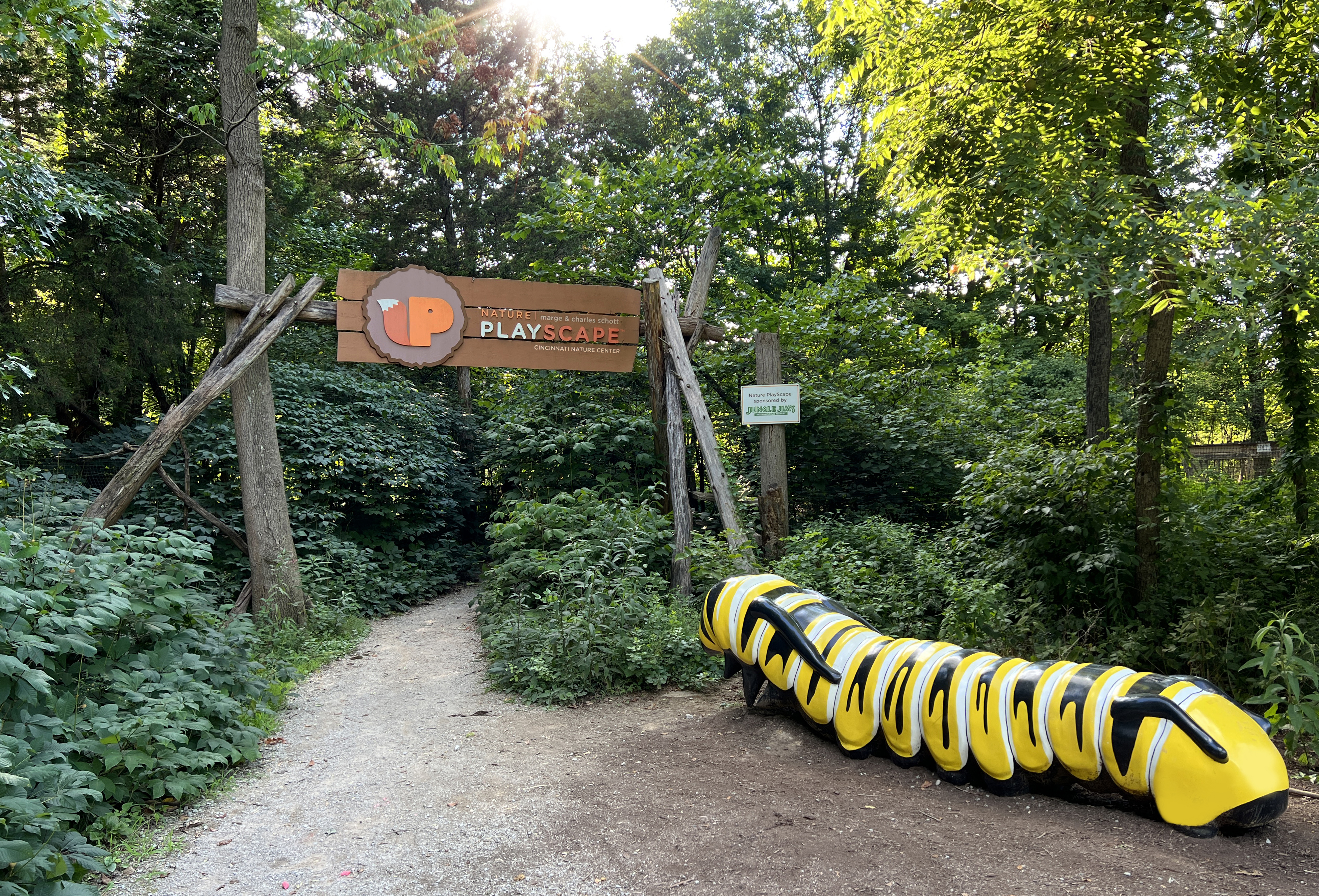 A graveled trail running under a sign that says "PlayScape". There is a large sculpture of a monarch caterpillar to the right. 